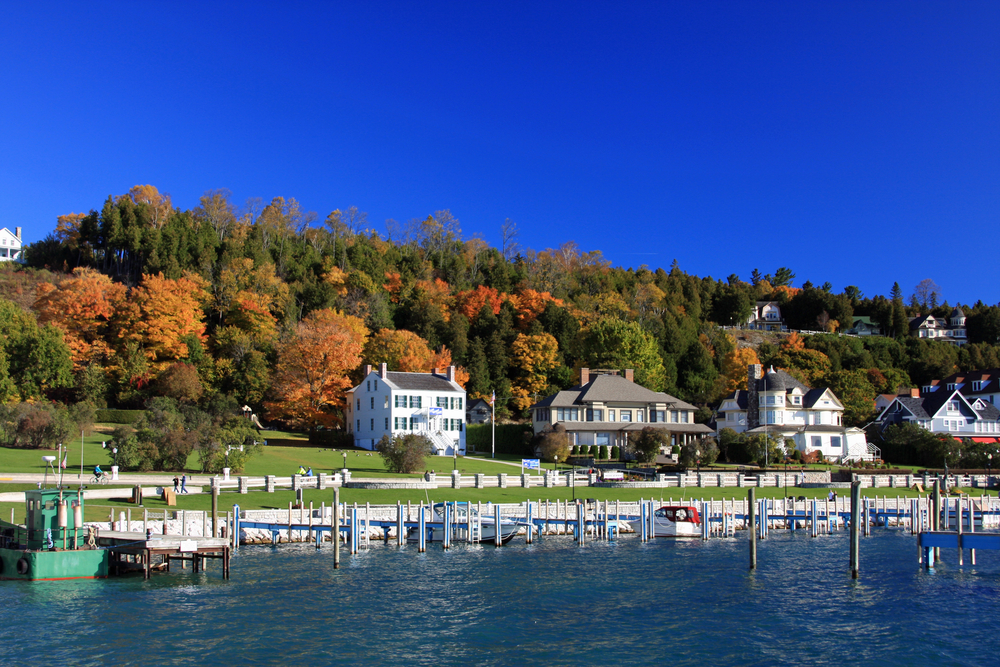 The view of houses along the shore of Mackinac Island in the fall. There are rows of docks for boats and trees with changing leaves. 