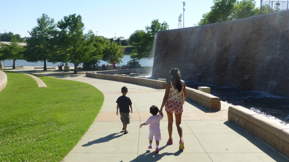 A woman walking in a park along the river with two small kids, one of the best things to do in Wichita. 