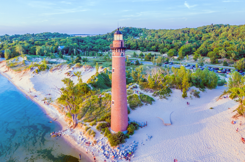 A red brick lighthouse on the edge of a lake surrounded by sandy dunes. The dunes also have trees, grasses, and shrubs growing on them. One of the best state parks in Michigan. 