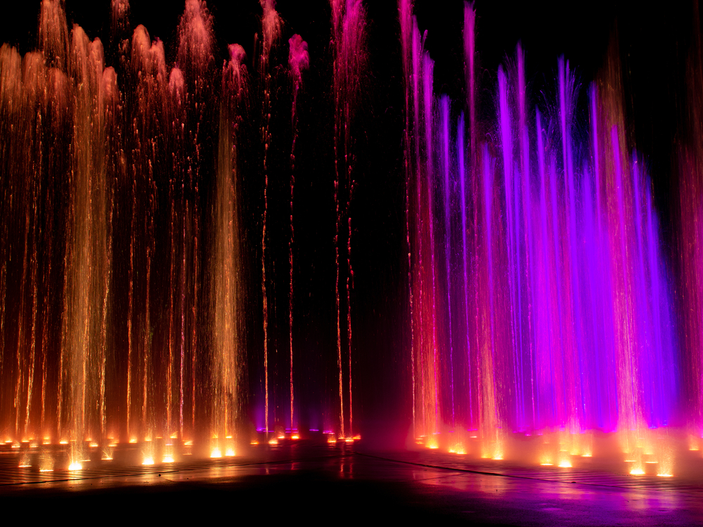 Large water fountains shooting out of the ground at night. The water streams are different colors and are part of a water fountain show. 