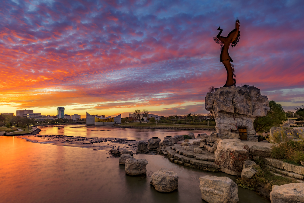 A sculpture of a Native American overlooking a pond and the city of Wichita at sunset. 