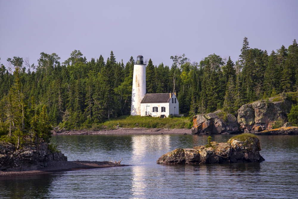 white lighthouse with red roof trees in the background and water with rocky outcroppings in forefront, at one of the islands in the Midwest