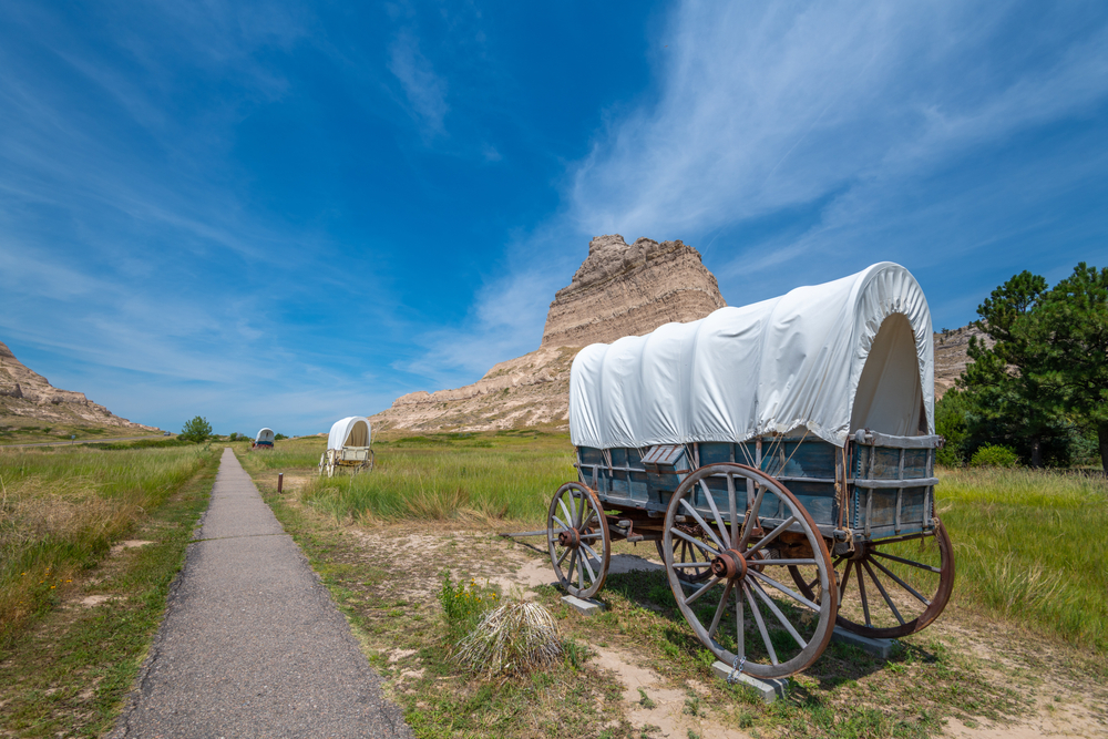 Covered wagons line the paved Oregon Trail in a prairie.