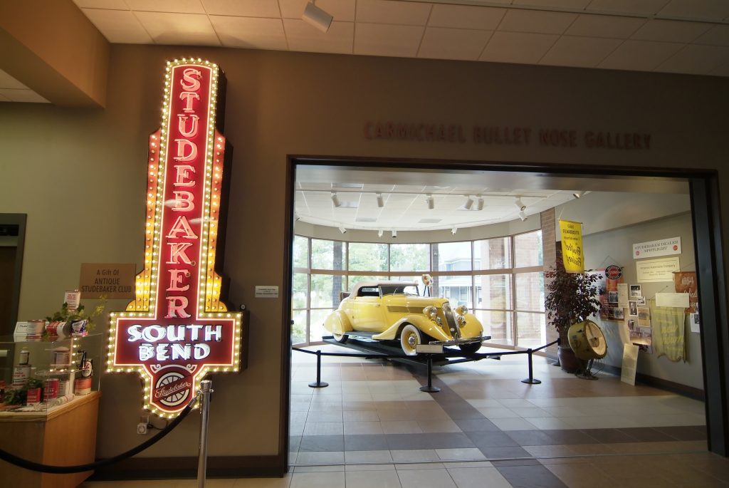 Inside the Studebaker National Museum with a neon Studebaker sign and a yellow car in the background.