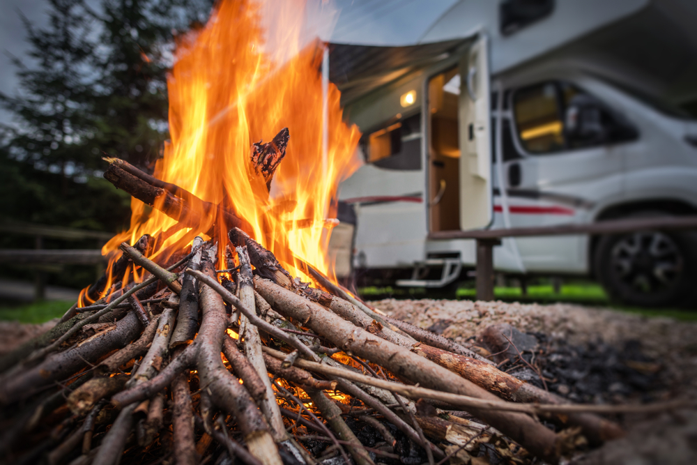 A closeup of a bonfire. Behind the bonfire is a small white RV with red and black stripes.