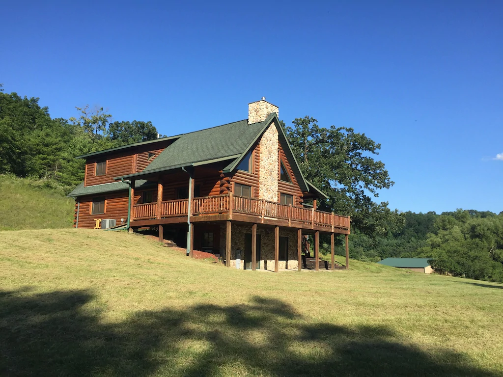 A large log cabin in a clearing with trees behind. a large stone chimney, and wooden railing surrounding back patio.. One of the coolest cabins in Wisconsin 