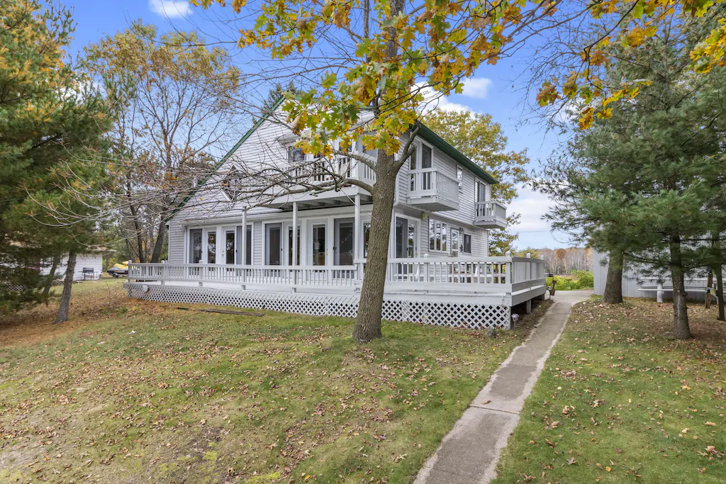 A white cabin with a large porch in a garden off a street 