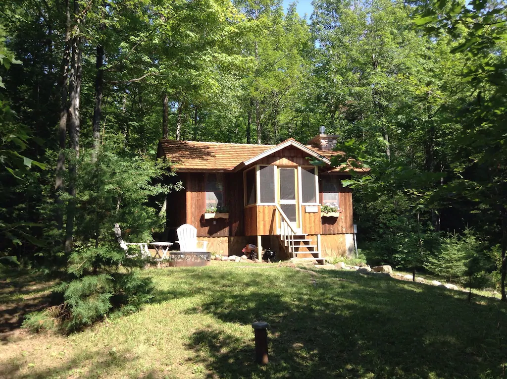 small brown log cabin with steps leading up to front door surrounded by trees. Fire pit and Adirondack chairs on left of this amazing Wisconsin cabin.