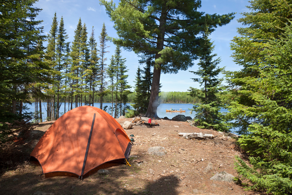 An orange tent on the side of a lake. You can see people kayaking in the lake in the distance. The campsite is surrounded by boulders and trees. 