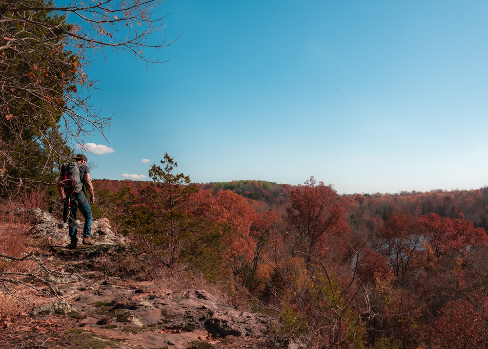 A person hiking on the side of a cliff on a rocky trail on a sunny day. You can see trees with red, orange, and green leaves on them. 