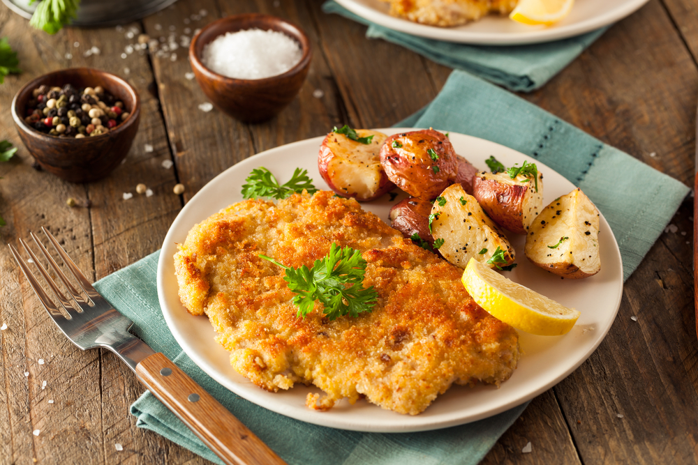 Pork Schnitzel on a plate with potatoes and parsley and a side of lemon on a  table with a napkin and fork. 