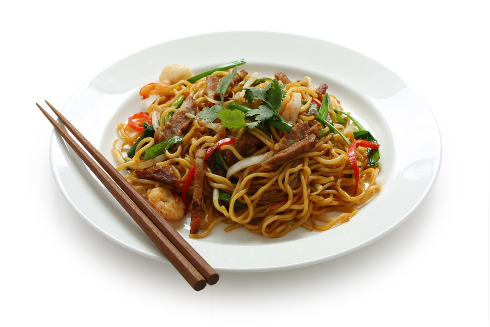 Brown noodles with veggies in white plate with wooden  chop sticks on left side of place.