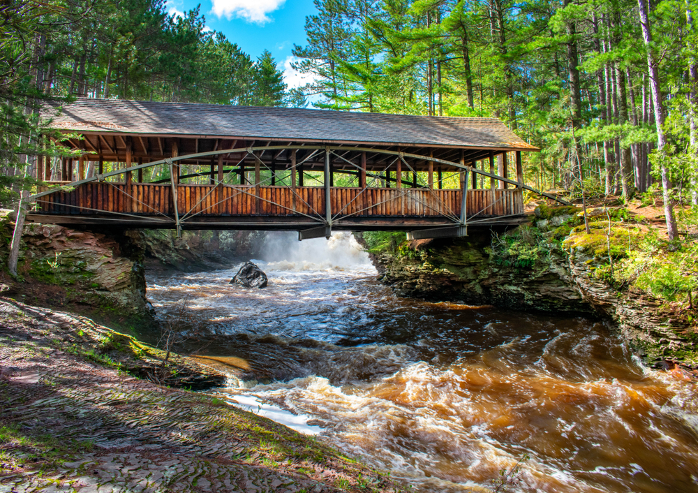 Wooden bridge with a roof crossing the river below. Trees are on either side of the bank. 