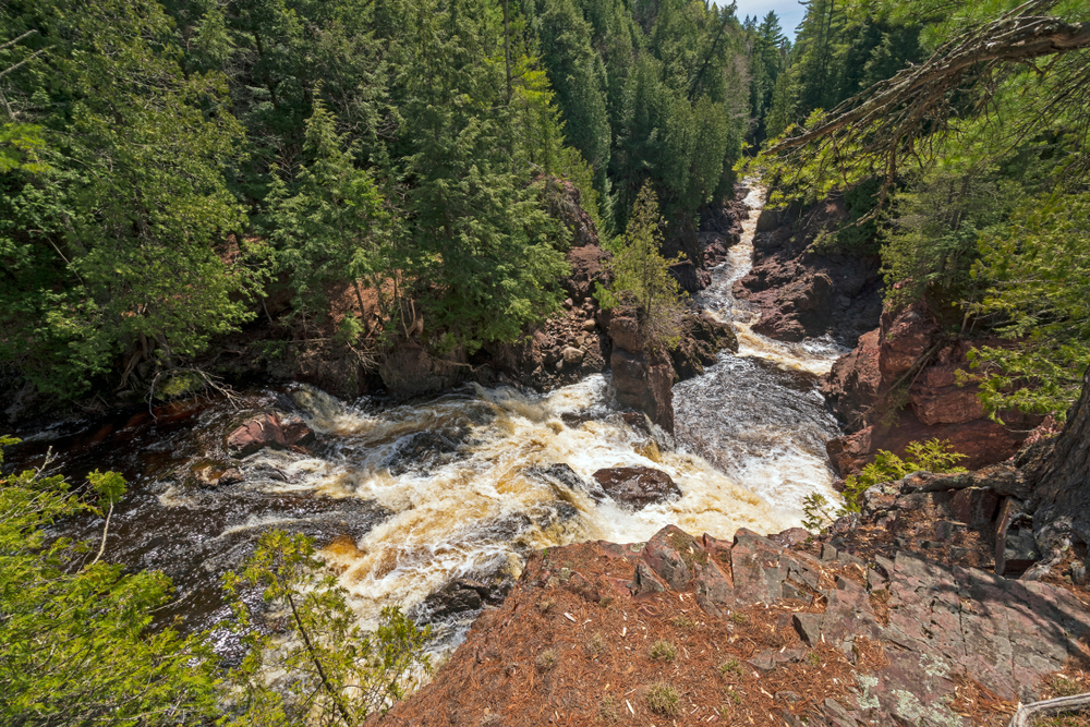 View from high of a ravine below with a river running through it. The river is falling down over the ricks forming cascades.  