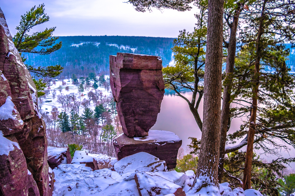 A large rock balanced on a smaller rock on the top of a cliff with a view to the valley below. Snow is on the ground. Devils Lake State Park is one of the best state parks in Wisconsin.