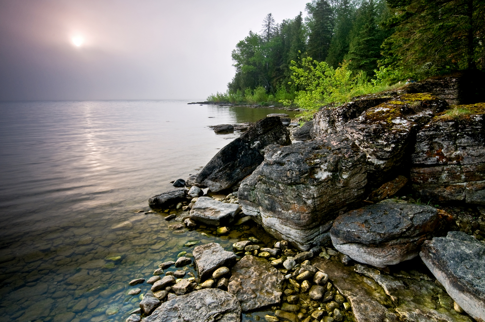 Rocky lake shoreline at dusk with trees in the background. 