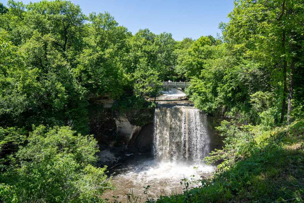 View looking down at Lower Minneopa Falls gushing into a gorge all surrounded by green trees.