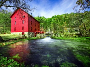 Red barn surrounded by gree trees and water in foreground in front of photo.