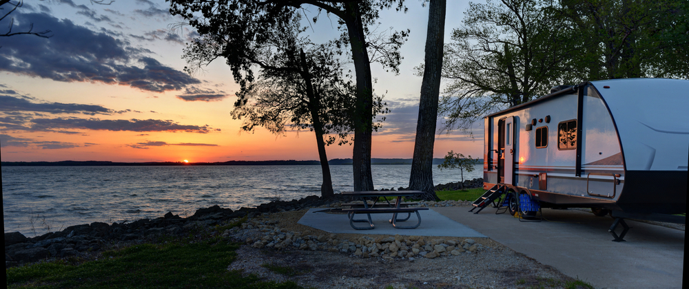 An RV on a cement slab with a picnic table. It is on the shore of a lake and the sun is setting. One of the best places for camping in Kansas.