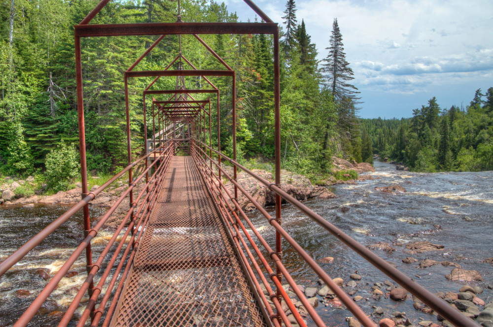 Red, metal bridge stretching across a rocky river into a forest for hiking in Minnesota.