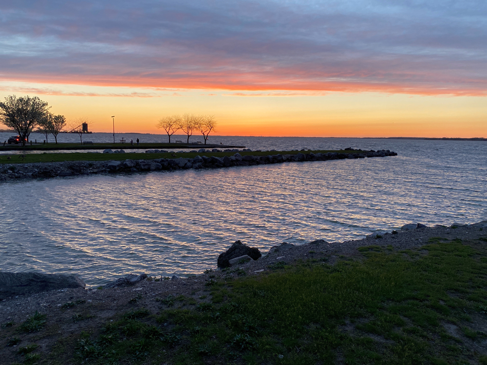 sunset from a shore with orange sunset sky and blue water in foreground and rocky break walls on Sandusky Beach.