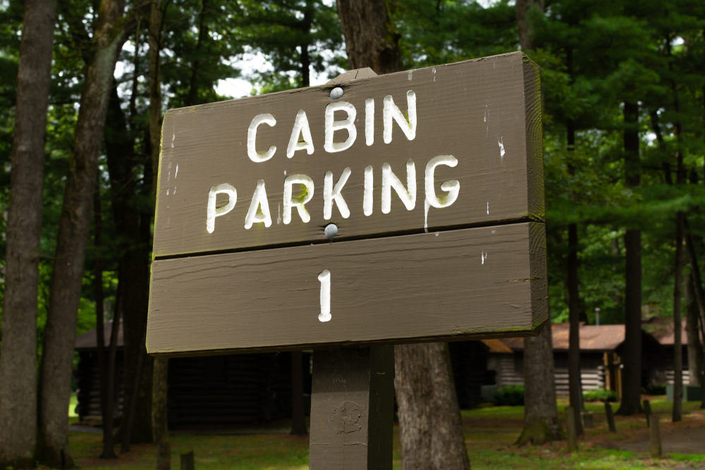 A wooden sign with white lettering that says 'cabin parking'. Behind it you can see trees and cabins in the distance. 