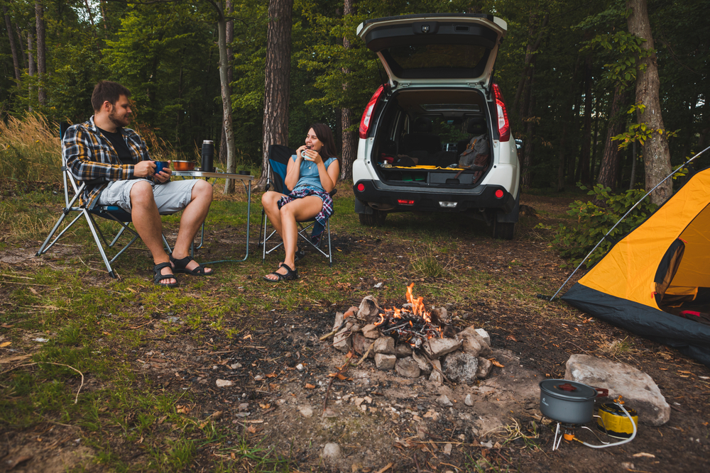 A couple sitting at a campsite where you can see a car that is open, a yellow tent, and a small fire pit. 