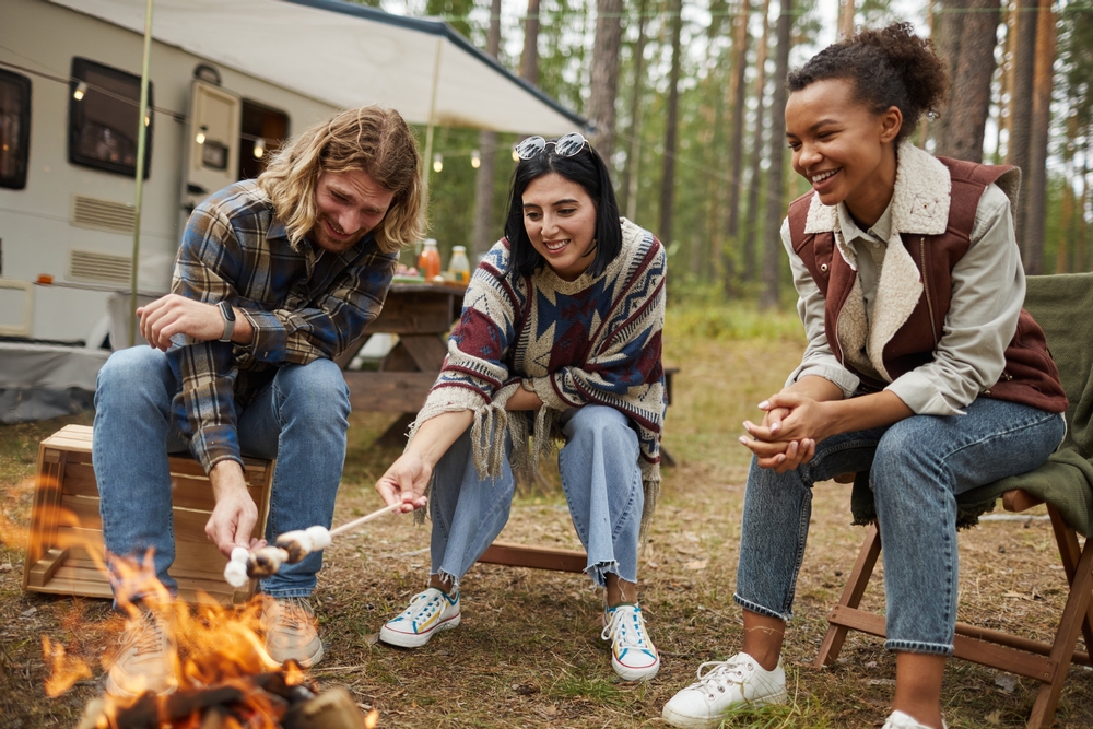 A group of people sitting around a bonfire roasting marshmallows with a camper in the background in the woods. 