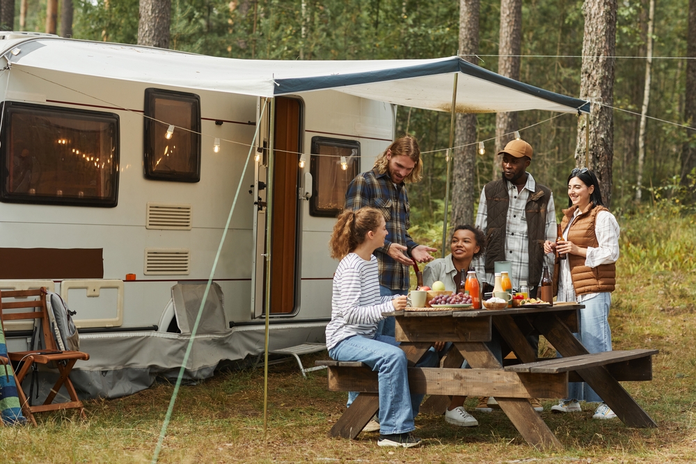A group of people hanging  out around an RV that has string lights and an awning. They are sitting at a picnic table eating. 