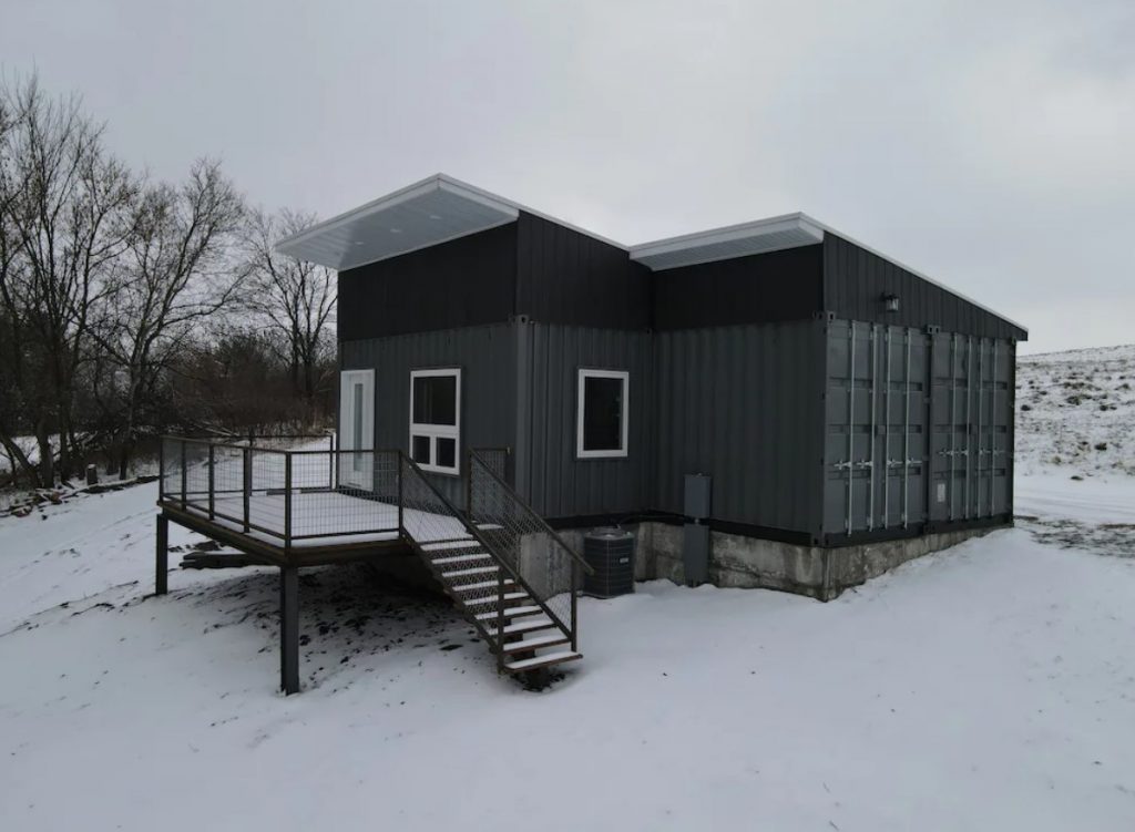 A small home made of grey and black shipping containers in a field covered in snow. At the front there is a small porch. One of the best places for glamping in Wisconsin. 