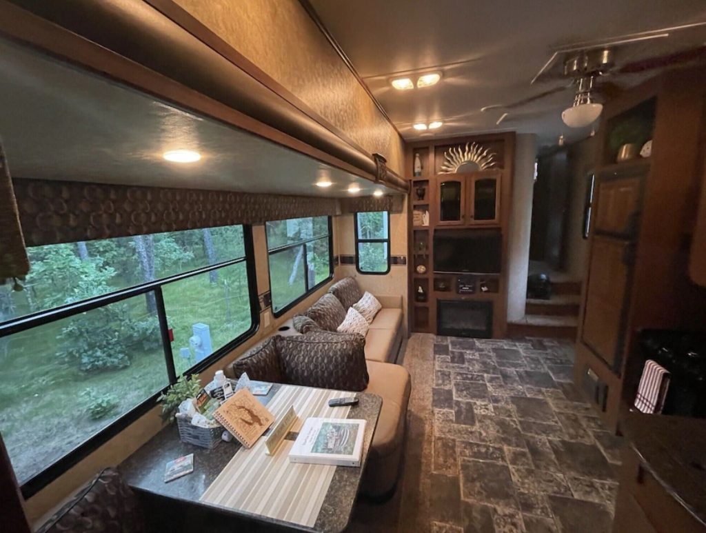 The interior of a luxury RV with a spacious living room that has a couch, dining table, lots of windows, and a small tv. 