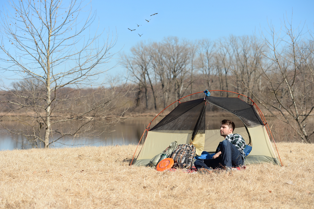 A man sitting outside of a brown and black tent that is next to a large body of water. The grass looks dead and there are no leaves on the trees. 