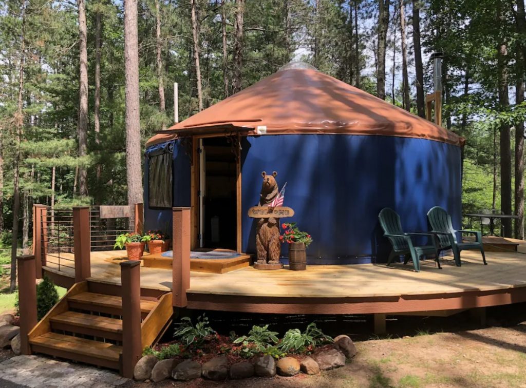 The exterior of a blue yurt with a brown roof. Around the yurt there is a deck with seating and it's in the woods. 