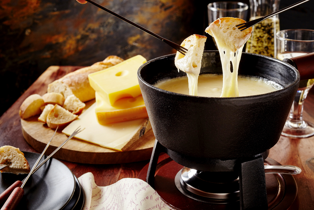 Cheese fondue on a wooden table with people dipping bread in. Their is bread and cheese on the table. 