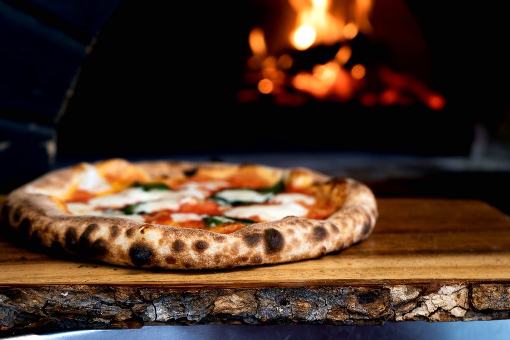 Wood fired hand made pizza with thick crust dressed with sauce and mozzarella cheese with fire oven in background.