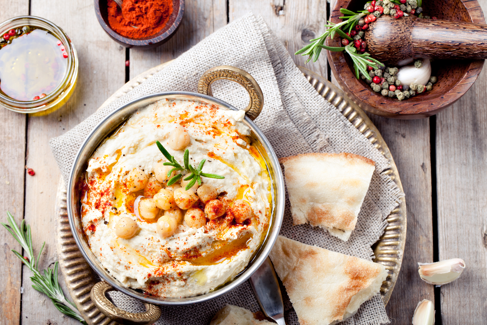 A dish of Hummus on a plate with flatbread. Chickpeas are on the top of the hummus and it is on a gold plate with a linen napkin. The artile is about restaurants in Oshkosh. 