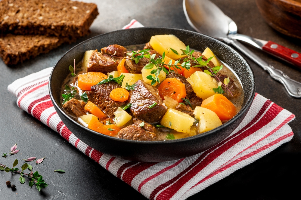 A bowl of Irish stew containing meat, carrots and potatoes on a red and white stripped napkin with cutlery off to the side.  Best food in Oshkosh