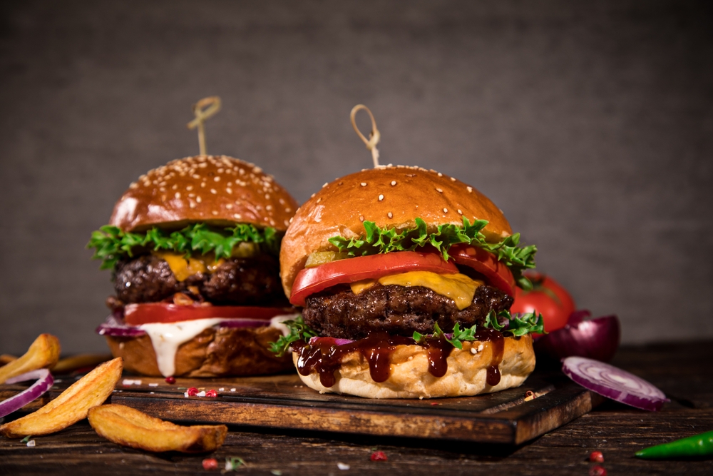 Two burgers in buns with barbecue sauce and cheese. It is on a wooden board with a few fries around. 