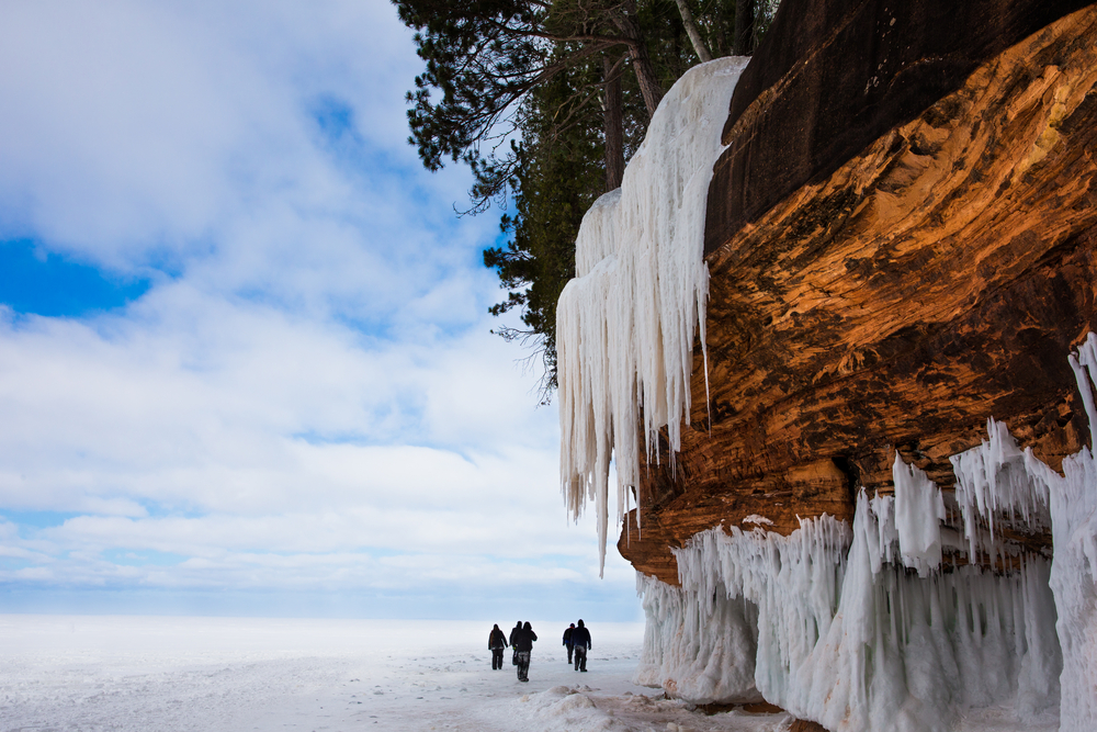 A cave rock formation with ice hanging from it. Snow is on the ground and there are people looking at the cave.  