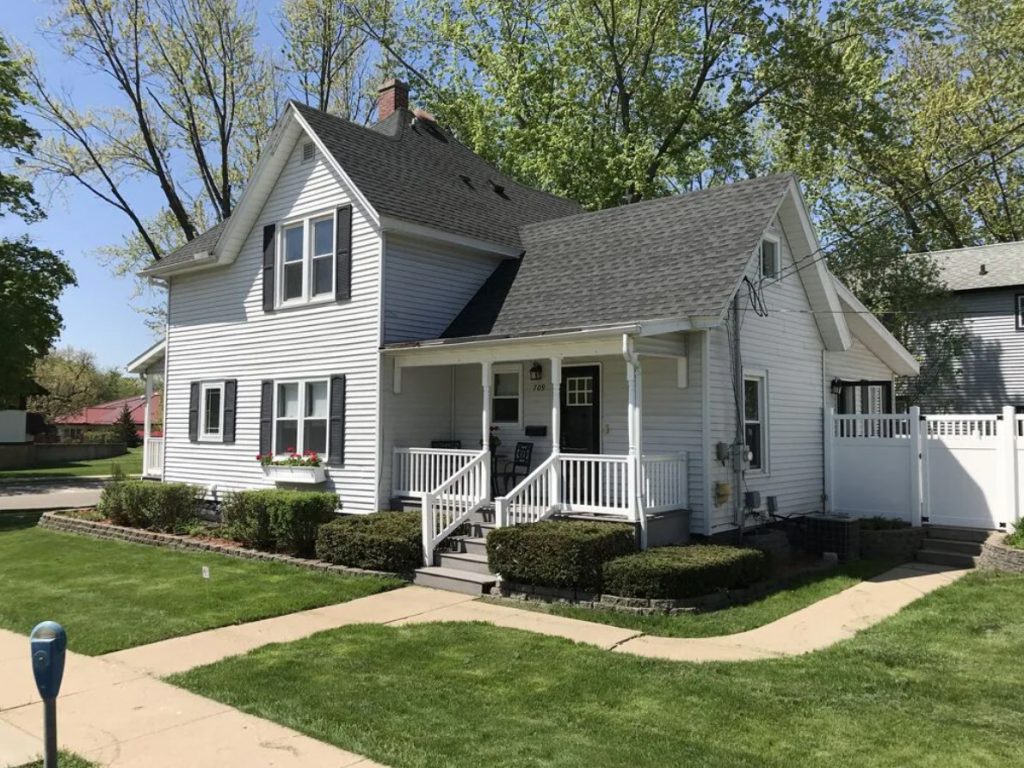A simple white house with a small front porch, a small green front yard, and a white fence along the side of it.  One of the cutest VRBOs in Wisconsin