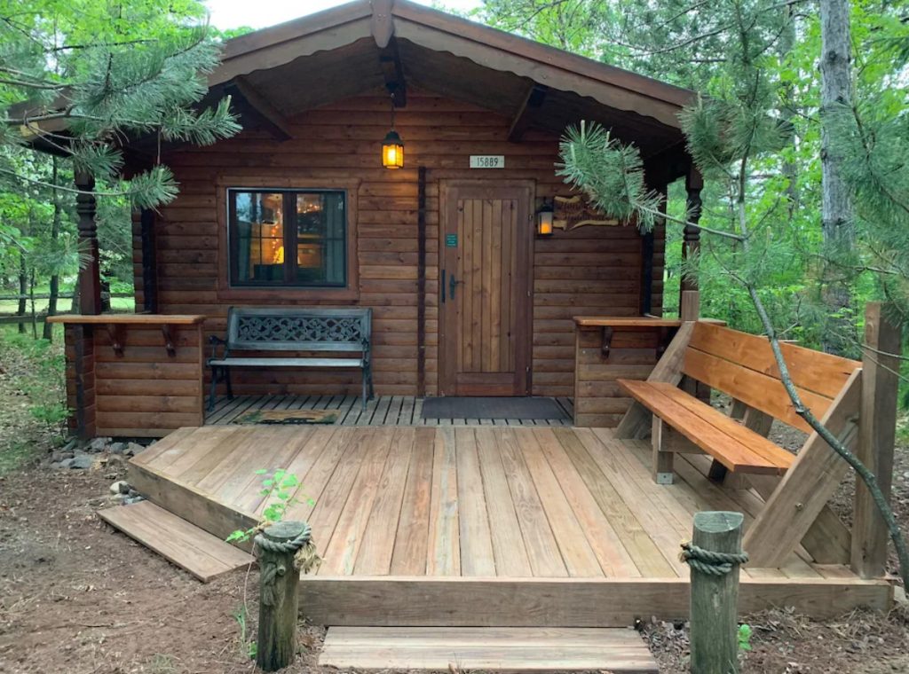The front of a small wood cabin with a deck that has two benches. It is surrounded by trees and is one of the best places for glamping in Wisconsin. 