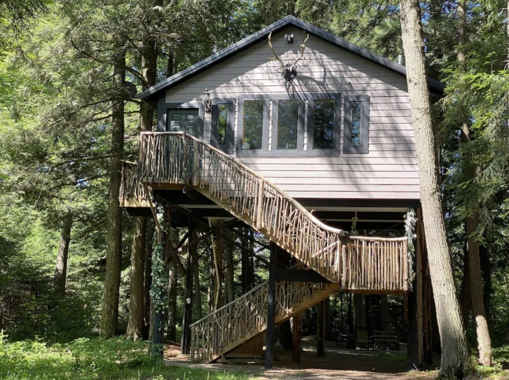 The exterior of a small grey treehouse perched among a group of trees. There is a staircase with a natural wood railing that leads you up to the house. It's one of the best places for glamping in Wisconsin.