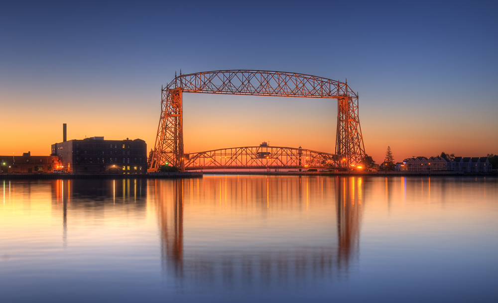 large metal bridge with yellow orange dawn sky and reflection in water of still lake.