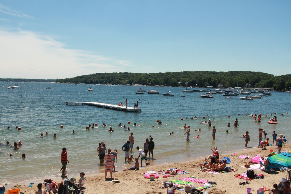 People on a beach with the lake and trees in the background. The beach is full of people  