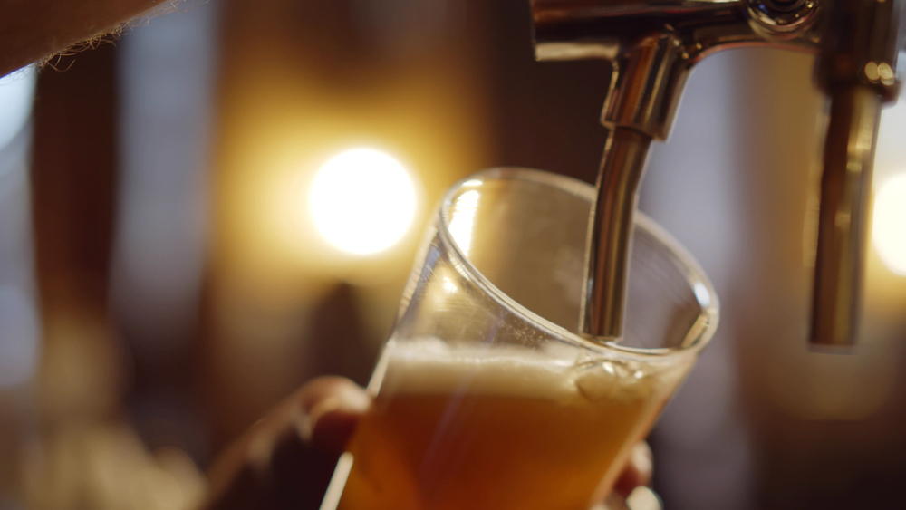 Beer been poured from a beer tap into  glass. A person is holding the glass.  