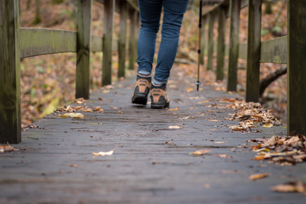 Picture of a girl wearing jeans and hiking boots walking along a bridge with leaves fallen on the floor. The picture is taken from behind. 