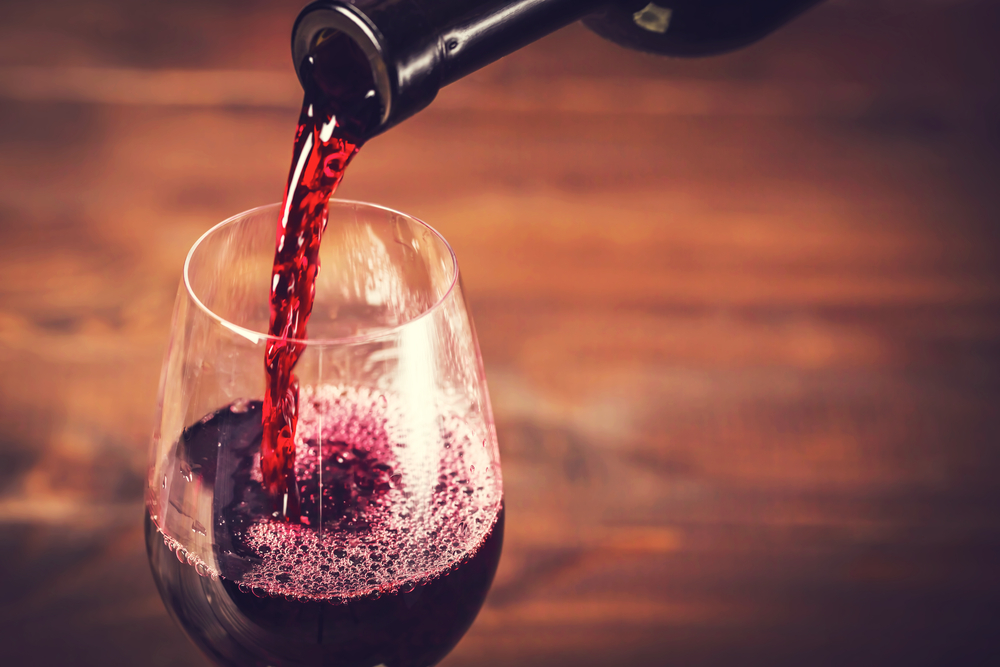 Red wine been poured from a bottle into a glass. The glass is on a wooden table. 