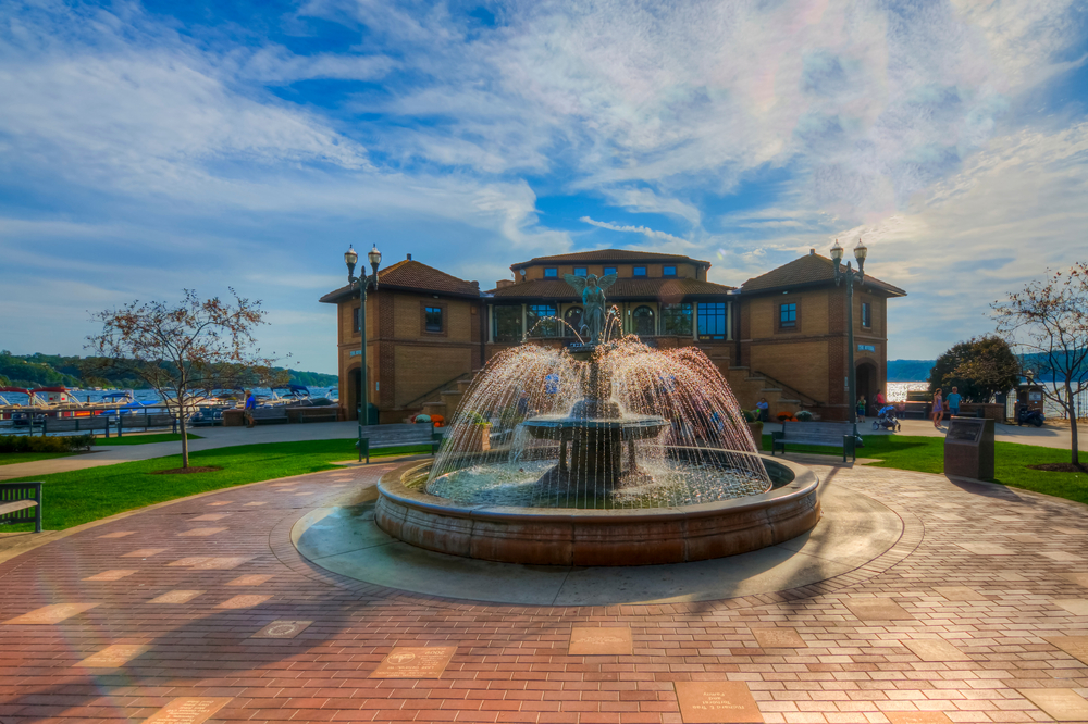 Fountain surrounded by a paved area and grass in a circle. It is in front of a building with a blue sky and water in the background.  In an article abut things to do in Lake Geneva. 