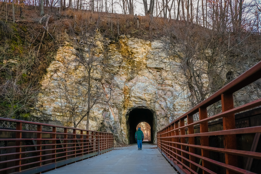 Winter view of bluff by Missouri River and tunnel through it build for old railroad converted into recreation trail at sunset; figure of man entering tunnel; bridge in foreground