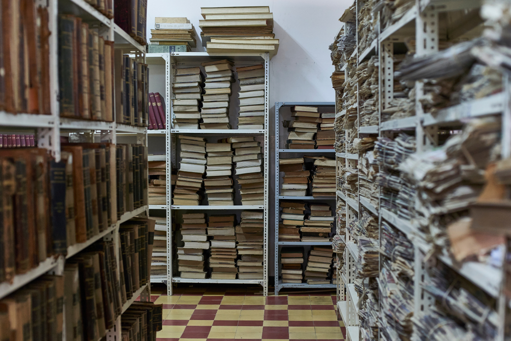 An archive room with shelves full of old books, newspapers, and other various items. 